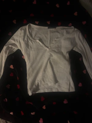 Vit långärmad topp med knappar - Vit långärmad topp med v-ringning och små knappar framtill. Tunn ribbad struktur och lätt croppad passform. Perfekt att styla med jeans eller kjol för en enkel och fräsch look. Den är insydd av mig (inte mitt bästa verk). 