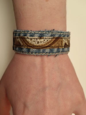 Brett armband i denim och tyg - Unikt armband i blå denim med brunt och beige tygband med mönster. Armbandet har råa kanter för en cool, avslappnad look och stängs med en svart tryckknapp i plast. Perfekt accessoar för dig som gillar en personlig stil. Ca 19 cm långt.
