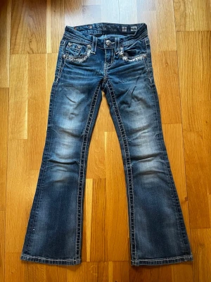 Mörkblå bootcut jeans från Miss Me - Snygga mörkblå bootcut jeans från Miss Me med slitningar och dekorativa detaljer på fickorna. Jeansen har kontrastsömmar, låg midja och broderade bakfickor med bling. Perfekta för dig som gillar en trendig och avslappnad stil. En knapp har dock gått av på backfickan och en av fickorna går inte att öppna helt.