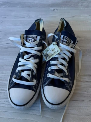 Converse Chuck Taylor All Star sneakers storlek 42 - Klassiska marinblå Converse All Star sneakers med lågt skaft, vita gummisulor och tåhättor. Skorna har vita snören och tydliga logotyper på plösen. Tillverkade i canvas med kontrastsömmar och metallöljetter. Perfekta för en avslappnad och cool stil.