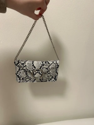 Kuvertväska med ormmönster och kedja - Säljer en snygg kuvertväska i svartvitt ormmönster med silverfärgad kedjerem. Väskan har ett stort runt spänne framtill och är tillverkad i konstläder. Perfekt accessoar för att lyfta din outfit.
