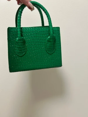 Grön minihandväska i krokopräglat material - Snygg grön minihandväska med två handtag och croco-mönstrad yta. Väskan har en boxig form och är tillverkad i konstläder med glansig finish. Perfekt accessoar för att lyfta din outfit med en poppig färg.