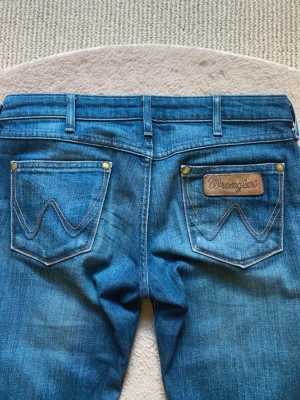 Vintage Wrangler lågmidjade jeans med detaljer - Snygga blå bootcut low waist jeans från vintage Wrangler med klassiska detaljer och logga på bakfickan. Jeansen har normal passform, fem fickor och är tillverkade i slitstarkt denim. Perfekta för dig som gillar retrovibbar och vill ha ett par jeans med lite utsvängda ben!😻Passar till varje plagg💋Inga defekter eller fel!! Innerben 80cm, midja 36cm, grenhöjd 19cm! Storlek W26 & L32, passar XS💕Billigare på min Vinted: 4annie1. Pris kan diskuteras❤️