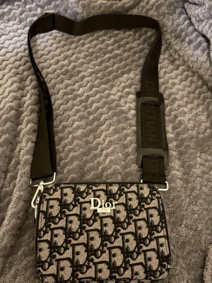 Dior axelväska med monogram - Snygg axelväska från Dior med svart och beige monogrammönster och bred svart axelrem där det står 'Christian Dior'. Väskan har silverfärgade detaljer och dragkedja upptill. Perfekt för dig som gillar lyxiga accessoarer och vill sticka ut.