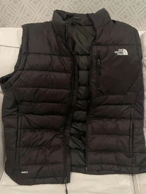 Svart dunväst från The North Face - Snygg svart dunväst från The North Face med hög krage och dragkedja framtill. Västen har en bröstficka med dragkedja och diskret logga på både fram- och baksidan. Tillverkad i slitstarkt material och fylld med dun för extra värme. Perfekt för lager-på-lager-stil. Helt oanvänd. Ny 1500kr