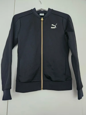 Svart ziptröja från Puma - Snygg svart ziptröja från Puma med guldfärgad dragkedja och logga på bröstet. Tröjan har ribbade muddar vid ärmslut och nederkant samt en klassisk sportig look. Perfekt till träning eller chill. Materialet är polyester.