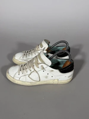 Philippe model skor - Säljer ett par vita sneakers med svart detalj på hälen och beige snören. Skorna har en klassisk låg siluett och är tillverkade i läder med gummisula. Perfekta för dig som gillar en clean och enkel stil.