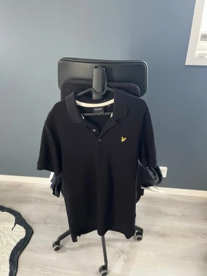 Svart pikétröja från Lyle & Scott - Klassisk svart pikétröja från Lyle & Scott med korta ärmar och gul broderad logga på bröstet. Tröjan har krage och knappar framtill, perfekt för en clean och stilren look.