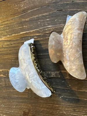 Två stora hårklämmor i pärlemor - Säljer två trendiga hårklämmor i pärlemor-look, en i ljus beige och en i vit. Klämmorna har en snygg vågig form och metallfäste. Perfekta för att hålla upp håret och ge en cool touch till din frisyr.