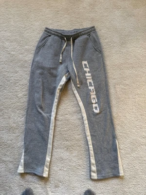 Grå mjukisbyxor med CHICAGO-tryck - Snygga grå mjukisbyxor från Manfinity med vitt CHICAGO-tryck på ena benet och vita ränder längs sidorna. Byxorna har dragsko i midjan och två fickor. Tillverkade i mjukt och skönt material, perfekta för chill dagar eller träning.