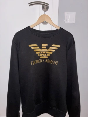 Svart sweatshirt från Giorgio Armani - Svart sweatshirt med stort guldfärgat Armani-tryck och logga på bröstet. Klassisk rund halsringning och ribbade muddar vid ärmslut och nederkant. Perfekt för dig som gillar stilrena och exklusiva plagg med tydlig branding.