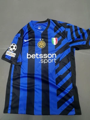 Inter Milan matchtröja blå/svart Nike - Snygg Inter Milan matchtröja i blått och svart med ränder, tillverkad av Nike. Tröjan har klubbmärke, sponsorlogga och Champions League-märke på ärmen. Tillverkad i lätt och ventilerande material, perfekt för fotboll eller träning.