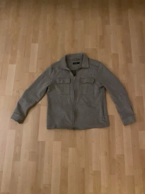 Grå overshirt med dragkedja - Stilren grå overshirt med dragkedja framtill och två bröstfickor med lock. Overshirten har klassisk krage och långa ärmar med knapp vid ärmslut. Perfekt lager på lager-plagg i mjukt material. Säljer även denna då den är för liten. Är 190 cm lång och bredaxlad.