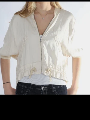 Beige vindjacka med dragkedja - Kort beige vindjacka med dragkedja framtill och snörning i midjan. Jackan har trekvartsärmar och en avslappnad passform. Perfekt för lager på lager och enkel att styla till olika outfits.