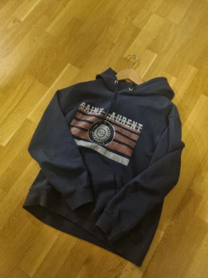 Mörkblå Saint Laurent hoodie - Snygg mörkblå hoodie från Saint Laurent med tryck framtill i vitt och rött samt texten 'SAINT LAURENT'. Klassisk huva med dragsko och ribbade muddar vid ärmar och nederkant. Perfekt för en avslappnad och trendig look.Storlek S. Väldigt snygg bra passform. 