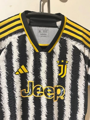 Juventus matchtröja Adidas svart/gul - Juventus hemmamatchtröja från säsongen 22/23. Tröjan har svartvitt zebramönster och gula detaljer. Tröjan har korta ärmar, rund halsringning och Juventus-logga samt Jeep-tryck på magen. Tillverkad i lätt och ventilerande material, perfekt för fotbollsträning. Tröjan är helt ny då jag bara dragit av prislappen. Säljer pga för stor 