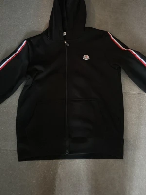 Svart Moncler hoodie storlek s - Snygg svart hoodie från Moncler med dragkedja framtill och huva. Ärmarna har röda, vita och blå ränder längs sidorna och Moncler-logga på bröstet. Perfekt för en sportig och trendig look. Mjuk och bekväm bomullsblandning.