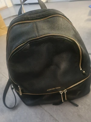 Svart ryggsäck från Michael Kors - Snygg svart ryggsäck i läder från Michael Kors med guldfärgade dragkedjor och logga framtill. Ryggsäcken har flera fack med dragkedja och justerbara axelband. Perfekt för dig som vill ha en stilren och praktisk väska till skolan eller stan.