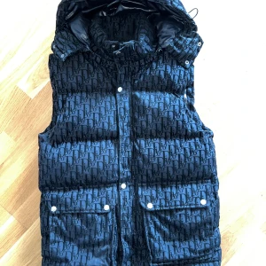 Svart dunväst från Dior med huva - Snygg svart dunväst från Dior med avtagbar huva och heltryckt logomönster. Västen har två stora fickor framtill med tryckknappar och hög krage. Mycket bra kvalité .