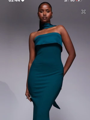 Mörk grön axelbandslös klänning - Säljer en elegant mörk grön klänning med axelbandslös design och lång, figurnära passform. Klänningen har en unik detalj med en bred halsduk som knyts runt halsen. Perfekt för dig som vill sticka ut med en modern och stilren look.