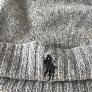 Grå stickad mössa från Polo Ralph Lauren - Snygg grå stickad mössa från Polo Ralph Lauren med klassisk ribbad kant och broderad svart logga framtill.