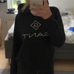 Svart hoodie från GANT  - Säljer en svart hoodie från GANT med stor vit logga och grafiskt tryck på bröstet. Klassisk huva och ribbade muddar vid ärmslut och nederkant. Perfekt för en chill och avslappnad stil. Tillverkad i mjukt material för extra komfort.