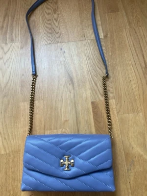 Ljuslila kuvertväska från Tory Burch - Säljer en ljuslila kuvertväska från Tory Burch i quiltat läder. Väskan har guldfärgad kedjerem och ikonisk logga framtill. Perfekt storlek för det viktigaste och ger en lyxig touch till din outfit.