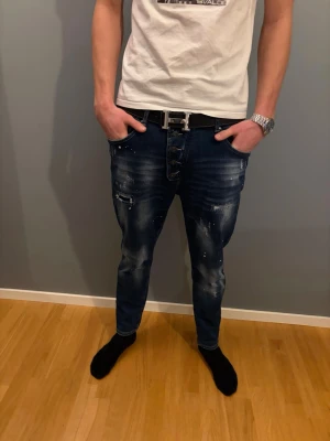 Mörkblå slim fit jeans med detaljer - Säljer ett par mörkblå slim fit jeans med slitningar och vita färgstänk för en cool, trendig look. Jeansen har knappgylf, dragkedjedetalj på ena benet och klassiska fem fickor. Perfekta för dig som gillar streetstyle och vill sticka ut.