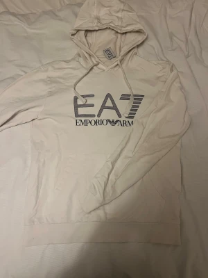 Vit hoodie från Emporio Armani EA7 - Snygg vit hoodie från Emporio Armani EA7 med stor logga framtill. Klassisk huva med dragsko och känguruficka. Tillverkad i mjukt material som känns skönt mot huden. Perfekt för dig som vill ha en stilren och trendig look.