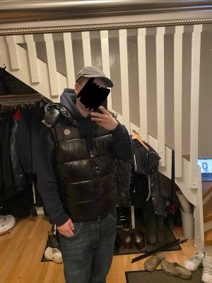 Svart dunväst från Moncler - Snygg svart dunväst från Moncler med glansig finish och klassisk logotyp på bröstet. Västen har hög krage och breda kanaler, perfekt att bära över en hoodie för en trendig look. HAR NFC SCAN! 