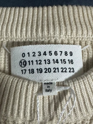 Beige stickad tröja Maison Margiela - Säljer en stilren beige stickad tröja från Maison Margiela. Tröjan har rund halsringning, ribbstickade muddar och är tillverkad i Italien. Perfekt för dig som gillar minimalistisk och exklusiv design.