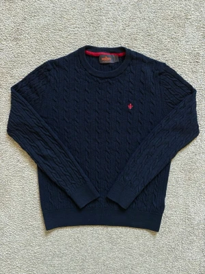 Mörkblå kabelstickad tröja Morris - Kabelstickad mörkblå tröja från Morris med rund halsringning och diskret röd logga broderad på bröstet. Tröjan har långa ärmar och ribbade muddar vid ärmslut och nederkant. Perfekt för dig som gillar klassisk och stilren design. Tröjan är i barnstorlek S vilket motsvarar storlek 152.