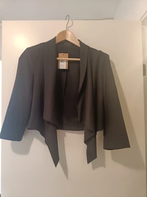 Svart kort kavaj med draperad front - Stilren svart kavaj med öppen, draperad front och trekvartsärmar. Modellen är kort och har en asymmetrisk nederkant som ger en modern look. Perfekt att slänga över en topp för en snygg outfit. Materialet känns mjukt och följsamt.