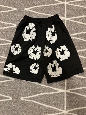 Svarta shorts med vita blommor - Säljer ett par svarta shorts med elastisk midja och stora vita blommönster över hela plagget. Shortsen har en avslappnad passform och är tillverkade i ett mjukt, lätt material som passar perfekt till sommaren. Snygg och trendig design för dig som vill sticka ut.