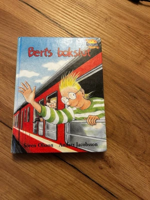 Berts bokslut av Olsson & Jacobsson - Rolig ungdomsbok med färgglad tecknad framsida där Bert vinkar från ett rött tåg. Boken är skriven av Sören Olsson och Anders Jacobsson och har hårda pärmar. Perfekt för dig som gillar humor och dagboksstil.