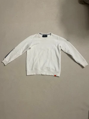 Vit sweatshirt från sailracing - Vit sweatshirt från sailracing med grå detaljer på armbågar och axlar. Tröjan har rund halsringning, ribbade muddar och en patch på ena ärmen. Materialet är mjukt och bekvämt, perfekt för en avslappnad stil.