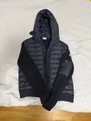 Mörkblå cardigan Moncler - Mörkblå pufferjacka från Moncler med stickade ärmar och quiltad huva. Jackan har dragkedja framtill och en liten ficka med dragkedja på ena ärmen. Perfekt för kalla dagar och snygg att matcha med jeans.