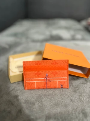  korthållare  - Snygg orange korthållare i tåligt läder med vit grafisk linjedesign och små illustrationer på båda sidor. Kommer med original ask och dustbag. Perfekt för dig som vill ha en färgstark accessoar från ett lyxigt märke.