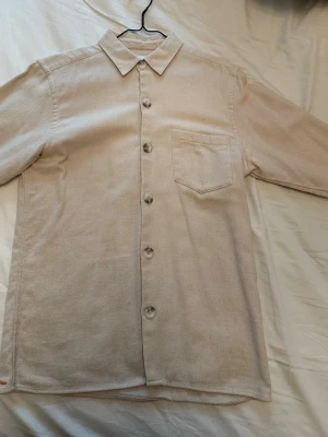 Beige overshirt från Ciszere i bomull med ficka - Stilren beige skjorta/overshirt från Ciszere i mjuk bomull med klassisk krage och bröstficka. Skjortan har långa ärmar och knäpps med stora knappar framtill. Perfekt för en clean och avslappnad look.
