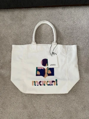 Isabel Marant Canvas Tote Bag - Snygg vit toteväska från Isabel Marant med broderad logga i lila, beige, grön och blått på framsidan. Väskan har två rejäla handtag och är gjord i kraftigt tyg, perfekt för att bära med sig allt man behöver. Märkets tagg sitter kvar.