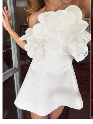 Vit axelbandslös kortklänning med blomdetaljer - Supersnygg vit axelbandslös kortklänning med stora 3D-blommor över bysten och axlarna. Klänningen har en utsvängd kjol och är perfekt för dig som vill sticka ut. Materialet ser ut att vara bomull eller polyester.