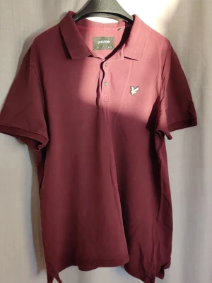Vinröd piké från Lyle & Scott XL - Säljer en vinröd pikétröja från Lyle & Scott med klassisk krage och knappslå framtill. Tröjan har korta ärmar och är tillverkad i mjuk bomull. På bröstet sitter den ikoniska broderade loggan i gult. Perfekt för en clean och stilren look.