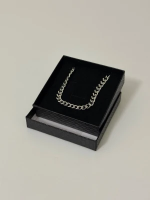 Silverfärgat kedjehalsband - Stilrent halsband i silverfärg med klassisk kedjedesign. Halsbandet har breda, platta länkar och mäter cirka 25 cm när det är vikt på mitten. Perfekt accessoar för en trendig look. Levereras i en svart presentask. 
