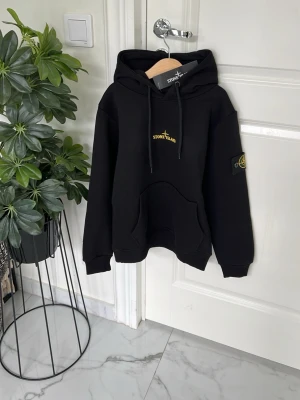 Svart hoodie från Stone Island barn - Snygg svart hoodie från Stone Island med gul logga på bröstet och stor tryckt logga på ryggen. Klassisk känguruficka framtill, huva med dragsko och ribbade muddar. Ikonisk patch på vänster ärm. Perfekt för en streetwear-look.
