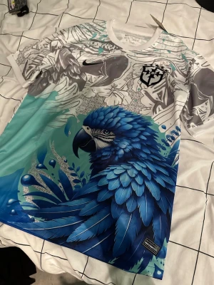 Nike t-shirt med blå papegoja - Unik t-shirt från Nike med stor blå papegoja och detaljerade grafiska mönster i vitt, grått och blått. T-shirten har korta ärmar och coola färgövergångar från vitt till blått. Perfekt för dig som vill sticka ut med en färgstark och kreativ look.