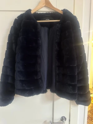 Svart fuskpälsjacka från Vero Moda - Snygg svart fuskpälsjacka från Vero Moda med mjukt, fluffigt material och horisontella sömmar. Jackan har rundad halsringning och är helfodrad för extra värme. Perfekt för dig som vill ha en trendig och mysig vinterlook.
