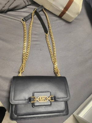 Svart axelväska Michael Kors - Snygg svart axelväska från Michael Kors i slätt läder med guldfärgade kedjor och MK-logga framtill. Väskan har en stilren, rektangulär form och detaljer i metall som ger en lyxig känsla. Perfekt för dig som vill ha en trendig och elegant accessoar.