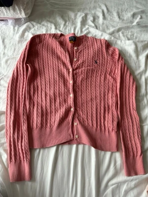 Rosa kabelstickad kofta Polo Ralph Lauren - Säljer en rosa kabelstickad kofta från Polo Ralph Lauren. Koftan har långa ärmar, knäppning framtill med pärlemorknappar och ribbade muddar. Klassisk broderad logga på bröstet. Perfekt för lager-på-lager och snygg till jeans. Xl i barne str tilsvarer en xs dame