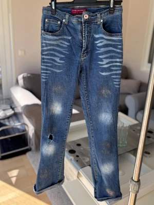 Blå jeans med zebramönster och glitter - Säljer ett par blå jeans från Angel Jeanswear med unikt zebramönster på framsidan. Jeansen har slitna detaljer, hål på ena benet och klassisk femficksdesign samt glitter. Materialet är denim.