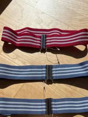 Randiga elastiska bälten i olika färger - Säljer tre breda elastiska bälten med randigt mönster och metallspänne, två i blått och ett i rött. Perfekt för att ge din outfit en cool touch och hålla allt på plats.